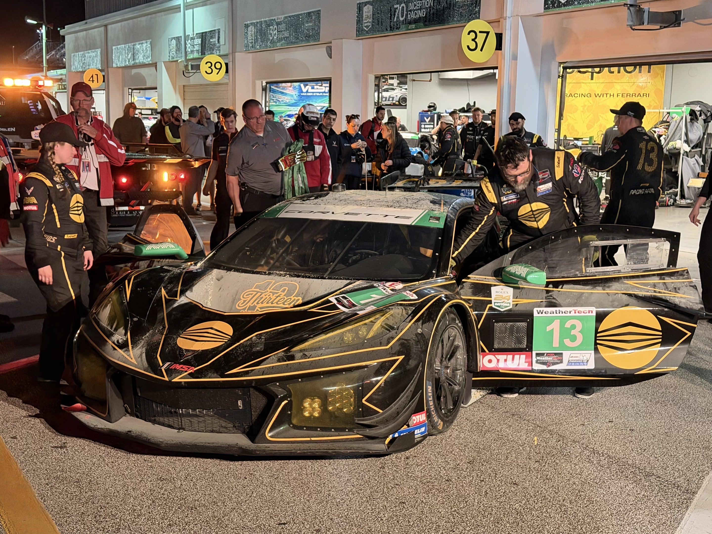 A crowd gathered around a burned up race car as the crew inspects it for safety and wheels it into the garage for repair
